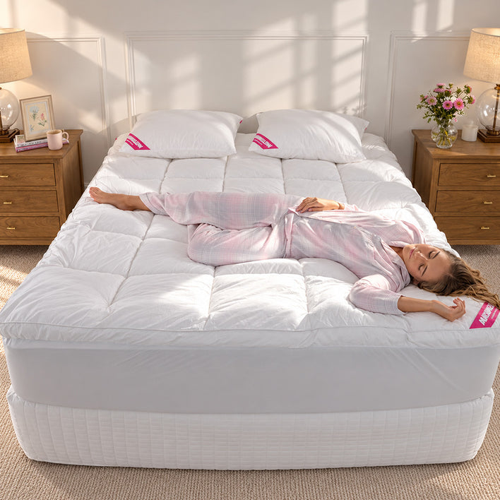 A woman in a pink pajama set lies on her back on a white quilted mattress topper with white pillows, in a brightly lit bedroom with wooden nightstands and lamps.