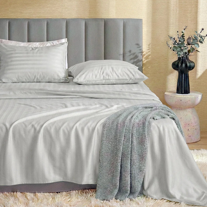 A neatly made bed features Super Sleeper Pro’s Buy 1 Get 1 FREE Royal Deluxe Imperial Dream Sheets™ Cotton Sateen 1200TC set in light gray stripes, with a textured gray blanket, upholstered headboard, and a side table with eucalyptus branches.