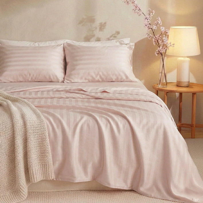 A bed is styled with Super Sleeper Pro's Buy 1 Get 1 FREE: Royal Deluxe Imperial Dream Sheets™ Cotton Sateen 1200TC 4pc Set + Free Organiser Bands, pale pink striped sheets and pillows, a cream blanket, and a nightstand with a beige lamp and pink flowers.
