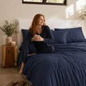 A woman with long red hair sits on a bed made with Super Sleeper Pro's Royal Deluxe Imperial Dream Sheets™ Cotton Sateen 1200TC 4pc Bedding Set, holding a mug, as natural light fills the room. A wooden nightstand beside her holds a plant and candle.