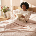 A woman sits in bed reading, wrapped in a light pink comforter over Super Sleeper Pro Royal Deluxe Imperial Dream Sheets™ Cotton Sateen 1200TC 4pc Bedding Set, with free organiser bands and a charming nightstand nearby.