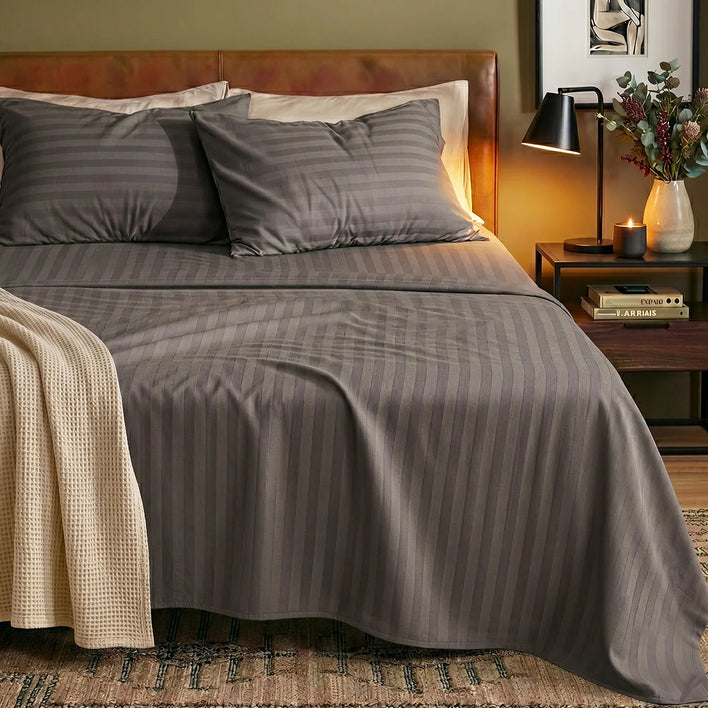 A bed made up with Super Sleeper Pro’s Royal Deluxe Imperial Dream Sheets™ Cotton Sateen 1200TC 4pc Bedding Set, topped with two pillows, sits beside a nightstand holding a lamp, candle, books, flowers, and a beige textured blanket.