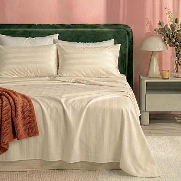 A bed dressed in Super Sleeper Pro's Royal Deluxe Imperial Dream Sheets™ Cotton Sateen 1200TC 4pc set, with two pillows and a rust throw, rests against a green headboard. A nightstand with lamp and vase sits to the right.