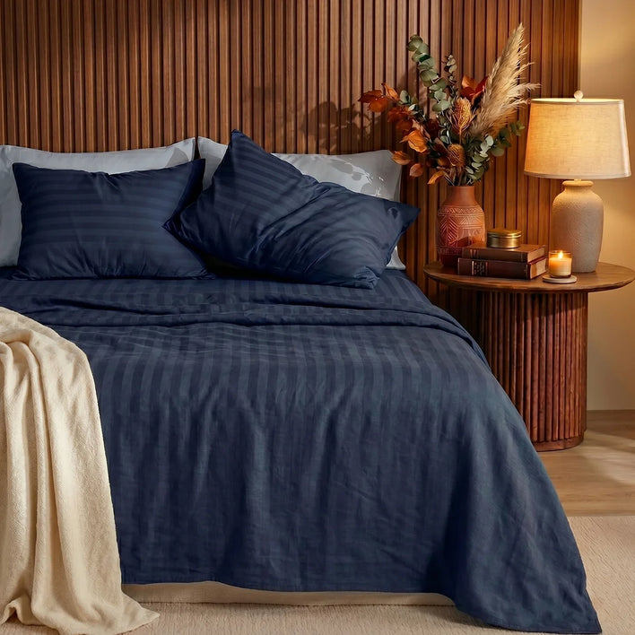 A neatly made bed features the Super Sleeper Pro Royal Deluxe Imperial Dream Sheets™ 1200TC Cotton Sateen 4pc Bedding Set. Two pillows, a beige blanket, and a round nightstand with a lamp, dried flowers, and candle complete the look against a slat wall.