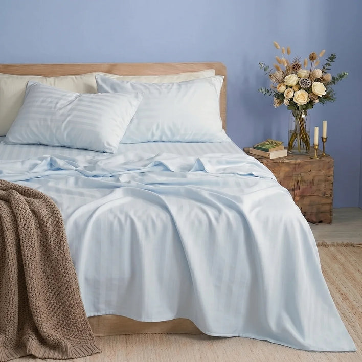 A made bed dressed in Super Sleeper Pro’s Royal Deluxe Imperial Dream Sheets™ Cotton Sateen 1200TC 4pc Set, in light blue stripes, with two pillows and a beige knit blanket at the foot, stands by a nightstand holding flowers, books, and candlesticks.