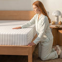A woman in striped pajamas kneels by a wooden bed, smoothing a Super Sleeper Pro Royal Deluxe Imperial Dream Sheets™ Cotton Sateen 1200TC 4pc Bedding Set with free organiser bands. A nightstand with a lamp, books, and decor is in the background.