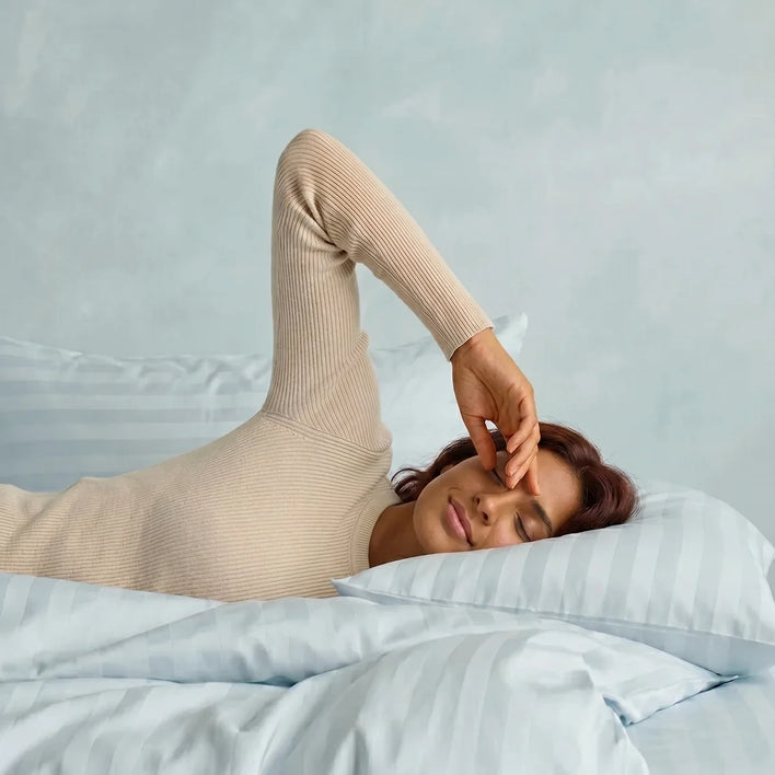 A person with long hair lies on a bed with Super Sleeper Pro Royal Deluxe Imperial Dream Sheets™ Cotton Sateen 1200TC 4pc Bedding Set, eyes closed, wearing a beige long-sleeve top. Free organiser bands included.