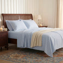 A neatly made bed with light blue striped Super Sleeper Pro Royal Deluxe Imperial Dream Sheets™ Cotton Sateen 1200TC 4pc Set, plus a cream knit blanket, between wooden nightstands holding lamps, photos, and candles in a white-paneled room with a patterned rug.
