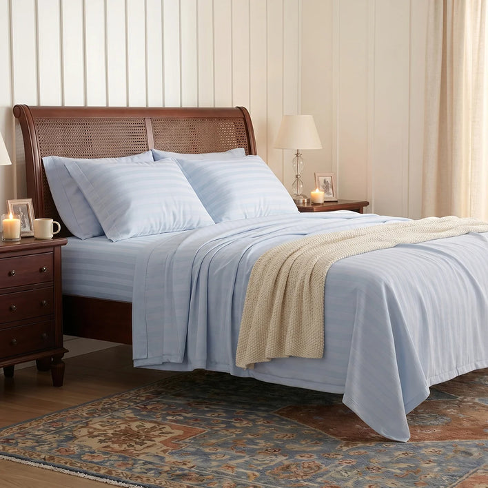 A neatly made bed with light blue striped Super Sleeper Pro Royal Deluxe Imperial Dream Sheets™ Cotton Sateen 1200TC 4pc Set, plus a cream knit blanket, between wooden nightstands holding lamps, photos, and candles in a white-paneled room with a patterned rug.