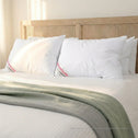 A neatly made bed with a rattan headboard features four Super Sleeper Pro Marshmellow™ Hotel Pillows and a white striped bedspread. A light green blanket is folded at the foot as sunlight streams onto this inviting scene.