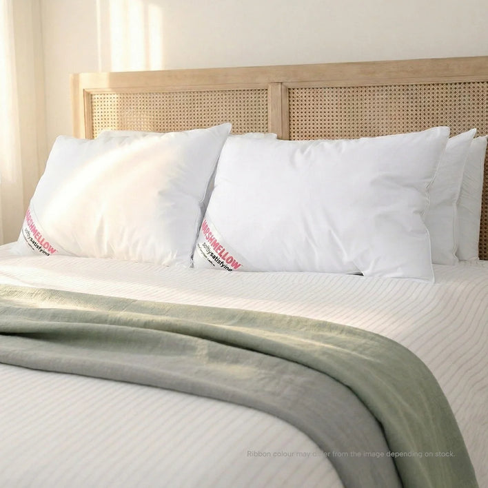 A neatly made bed with a rattan headboard features four Super Sleeper Pro Marshmellow™ Hotel Pillows and a white striped bedspread. A light green blanket is folded at the foot as sunlight streams onto this inviting scene.