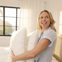 A woman smiles in a sunlit bedroom, holding two Super Sleeper Pro Marshmellow™ Hotel Pillows—ultra-soft, hotel-quality pillows designed for better sleep—while sunlight streams through a black-framed window behind her.