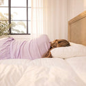 A person lies on a bed with white bedding, resting their head on a Super Sleeper Pro Marshmellow™ Hotel Pillow—an ultra-soft, luxury hotel-quality pillow for better sleep. Sunlight streams in through large windows with sheer white curtains.