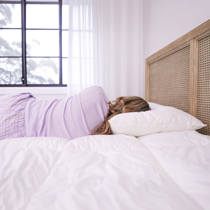 A person with long hair, wearing a lavender top and plaid pajama pants, lies on white bedding near a wooden headboard and window, resting on Super Sleeper Pro Marshmellow™ Hotel Pillows—ultra-soft, luxury hotel-quality pillows for better sleep.