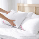 A person places a Super Sleeper Pro Marshmellow™ Hotel Pillow—an ultra-soft luxury hotel-quality pillow—onto a bed with similar pillows in a bright room with a light wooden headboard and crisp white bedding.