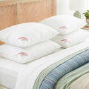 Four Super Sleeper Pro Marshmellow™ Hotel Pillows, ultra-soft and filled with 3D hollow fibre for a cool, luxury sleep, are stacked on a bed with white sheets and a green-blue blanket. A lamp and plant sit on the bedside table.