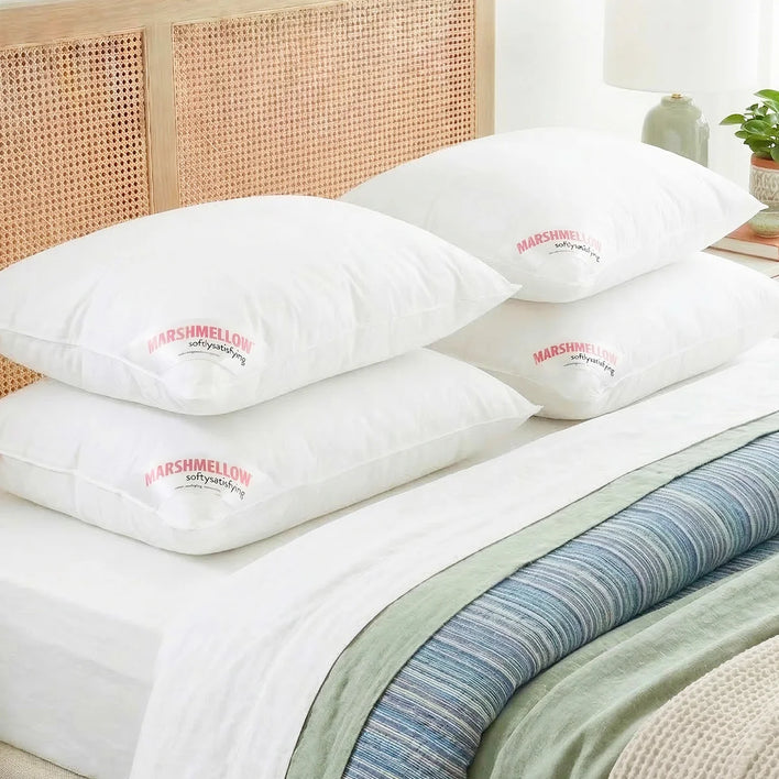 Four Super Sleeper Pro Marshmellow™ Hotel Pillows, ultra-soft and filled with 3D hollow fibre for a cool, luxury sleep, are stacked on a bed with white sheets and a green-blue blanket. A lamp and plant sit on the bedside table.