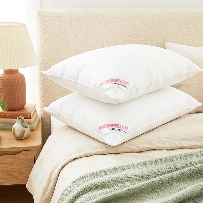 Two white Marshmellow™ Hotel Pillows by Super Sleeper Pro are stacked on a neatly made cream bed. On the bedside table, there’s a textured lamp and a small plant in a terracotta pot.