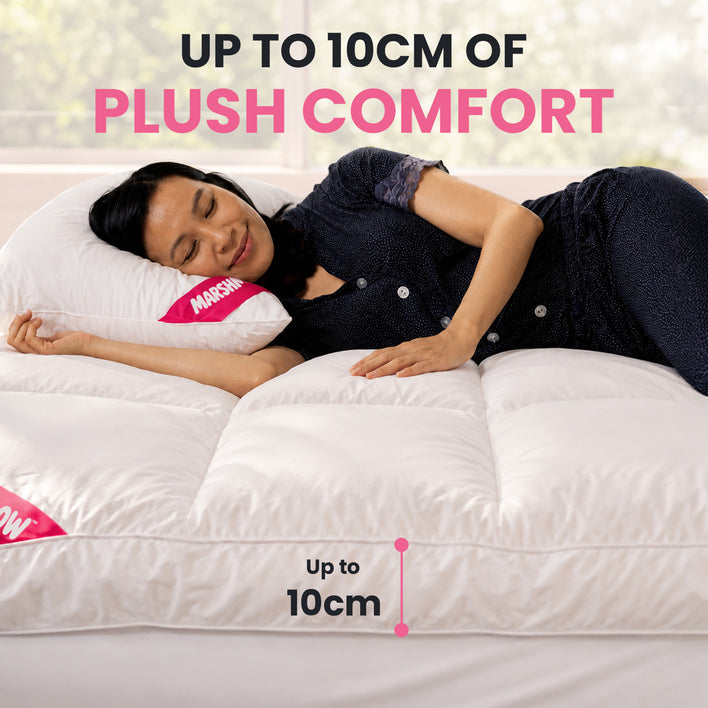 A woman lies on a white quilted mattress topper or bed pad in a bright bedroom setting, with promotional text reading 'UP TO 10CM OF PLUSH COMFORT' and a size callout indicating up to 10cm thickness.