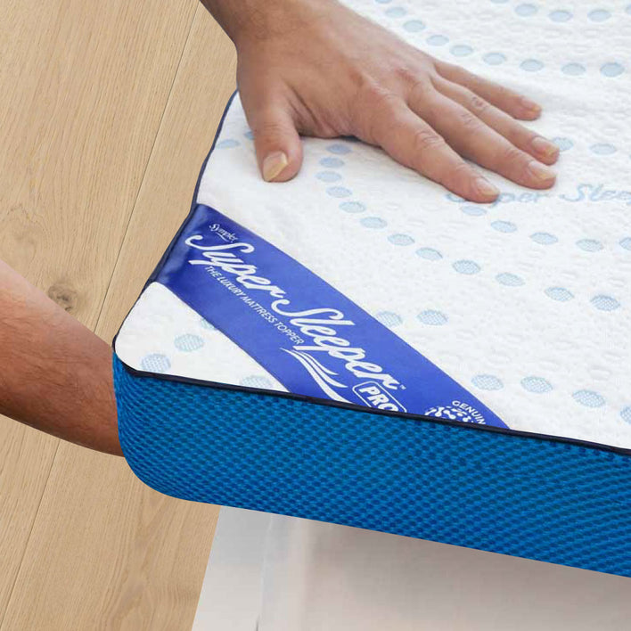 A hand presses on the corner of a Super Sleeper Pro bed featuring the #1 Customer Choice Straight 2 Sleep® Mattress Topper with Cooling Technology and bonus free pillows, all resting on a light wood floor.