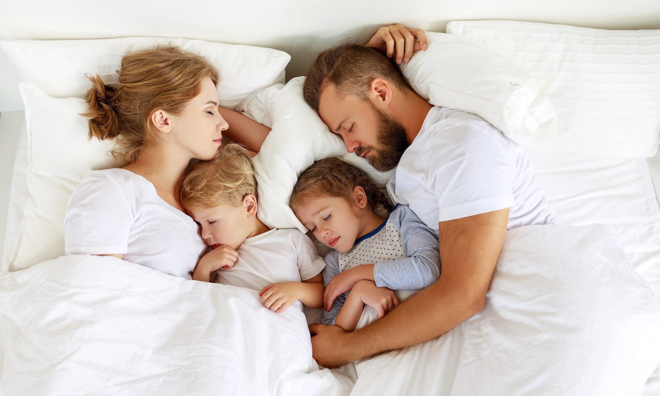 A man, woman, and two young children are lying together asleep in a bed, covered by a white blanket. All are wearing light-colored sleeping clothes and resting close to each other on white pillows.