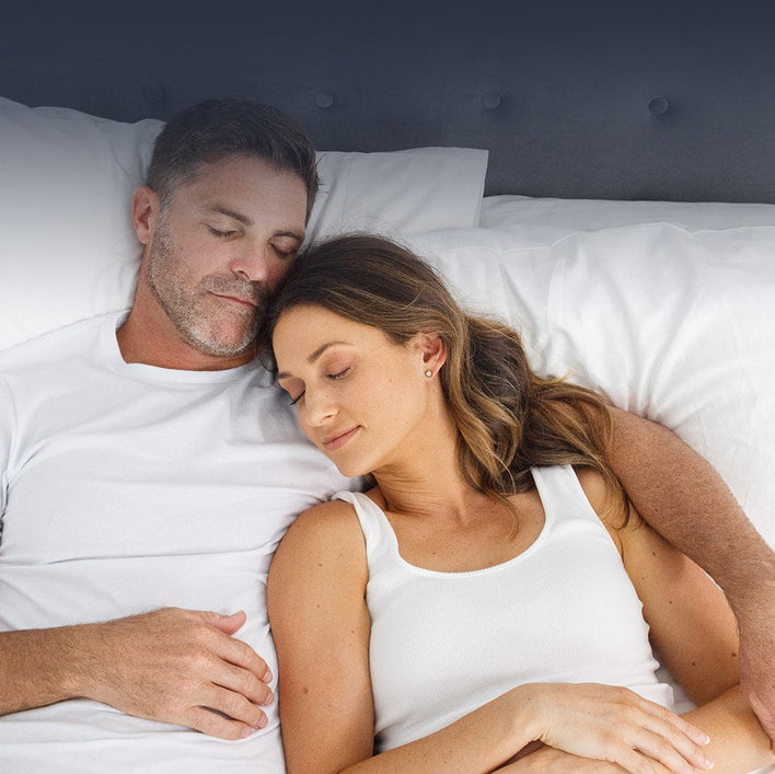 A man and woman, both in white tops, sleep peacefully together on the Super Sleeper Pro #1 Customer Choice Straight 2 Sleep® Mattress Topper with Cooling Technology and free pillows, their heads close on soft white pillows.