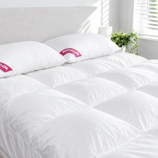A neatly made bed with a white comforter and two pillows is set against a light headboard, featuring the Super Sleeper Pro Marshmellow™ Plush Mattress Topper beside a window with white blinds and a green potted plant.