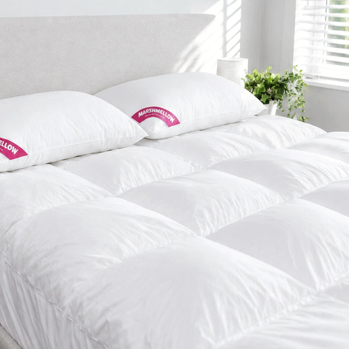 A neatly made bed with a white comforter and two pillows is set against a light headboard, featuring the Super Sleeper Pro Marshmellow™ Plush Mattress Topper beside a window with white blinds and a green potted plant.