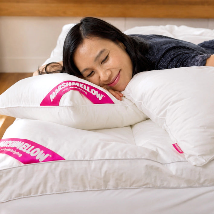 A woman with long dark hair lies on a bed, smiling with her eyes closed, enjoying the comfort of the Super Sleeper Pro Marshmellow™ Plush Mattress Topper beneath soft white bedding and pillows.
