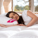 A woman with long brown hair sleeps on a bed, resting her head on a white pillow and lying on the Super Sleeper Pro Marshmellow™ Plush Mattress Topper, with sunlight streaming through large windows in the background.