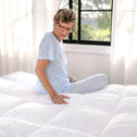 An older woman in light blue pajamas sits on a white comforter atop the Marshmellow™ Plush Mattress Topper by Super Sleeper Pro, gently touching the bedding. White curtains and a window revealing lush greenery complete the cozy scene.