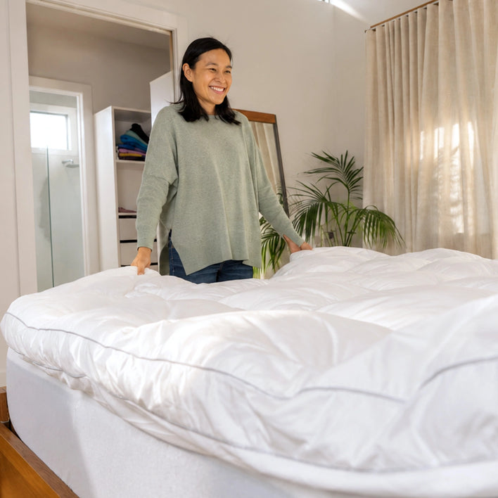 A woman smiles as she adjusts a white comforter over the Super Sleeper Pro Marshmellow™ Plush Mattress Topper. The bedroom features neutral walls, large curtains, a plant, and an open closet door in the background.