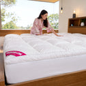 A woman in a pink robe smooths a large, white, quilted Super Sleeper Pro Marshmellow™ Plush Mattress Topper on a bed in a modern, sunlit bedroom with wooden furniture and large windows.