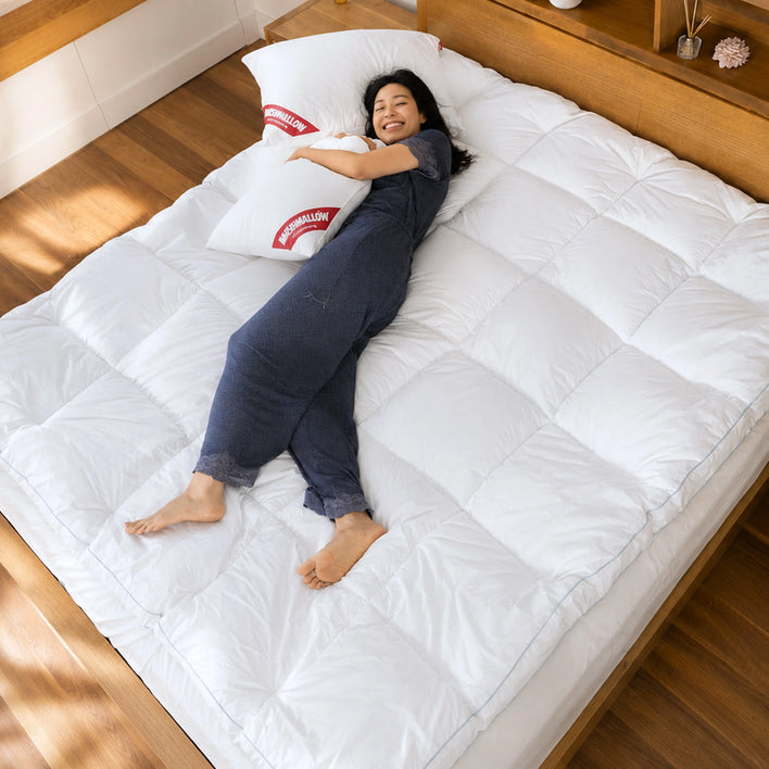 A person in dark pajamas lies on a large bed with a white comforter and Super Sleeper Pro Marshmellow™ Plush Mattress Topper, hugging two white pillows with red labels and smiling. The room has wooden floors and a wooden headboard with decor.