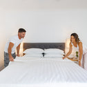 A man and women helping each other make their bed with luxury white sheets in a well-lit bedroom.