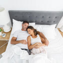 A man and a woman snuggled up together in a brightly lit bedroom, with luxury white bed linens enveloping them.