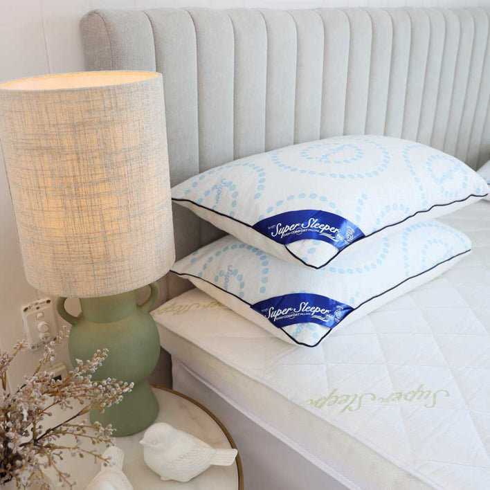 A bedside table with a green lamp and white decorative birds stands next to a bed with a tufted headboard. The Super Sleeper Pro Best Selling Every Comfort Pillow With Cooling Technology is stacked on the white quilted mattress for restful sleep.