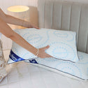 A plump pillow is resting on a bed with a grey headboard and a lamp on the side. A woman with a silver bracelet picks up an additional pillow