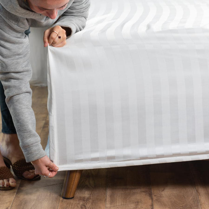 A woman bending over to fit a soft, white bedsheet onto the corner of her mattress.