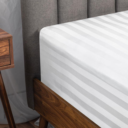 Left corner of a mattress which is made with a soft, white fitted bed sheet, placed on a dark wooden bed frame.