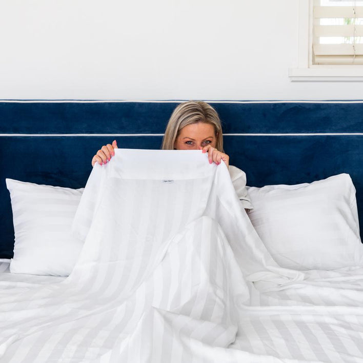 A light-haired lady sitting upright on her bed, adorned by luxury, white bed sheets.